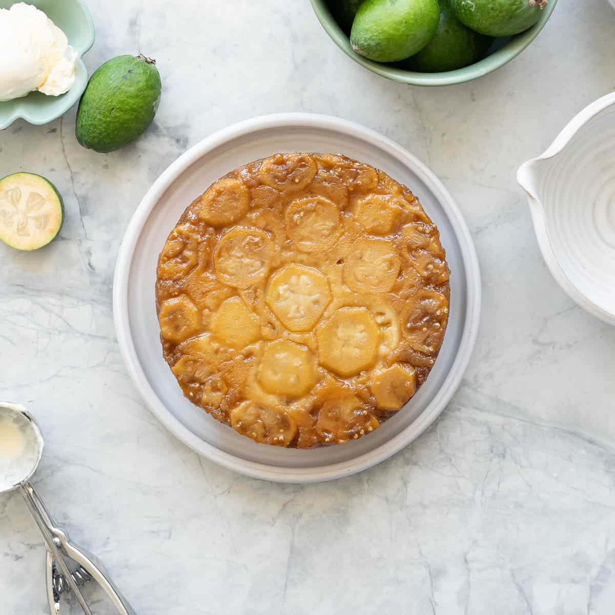 A feijoa cake on a ceramic plate resting on the bench next to a bowl of feijoas, a ramekin or ice cream and some halved feijoas.