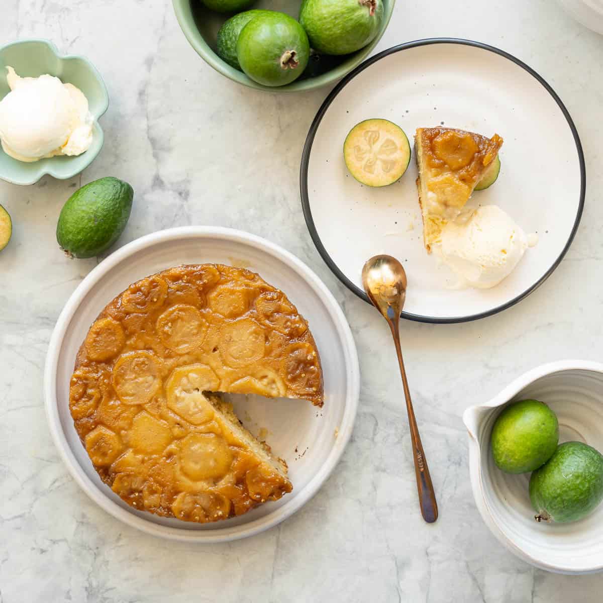 A feijoa cake resting on a ceramic plate on the bench next to one slice of the cake, a serving of ice cream and a brass spoon. which is all sitting next to whole and sliced feijoas.
