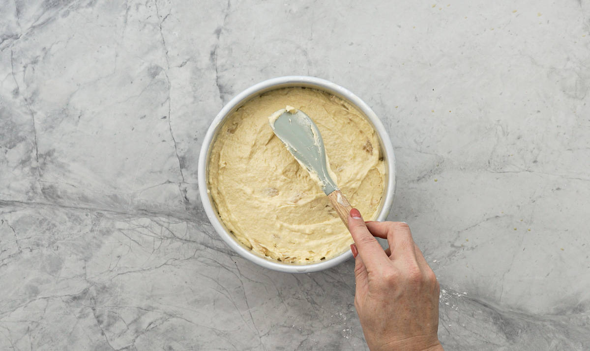 A hand spreading the feijoa cake batter over the feijoa caramel layer in the cake tin which is sitting on the bench.