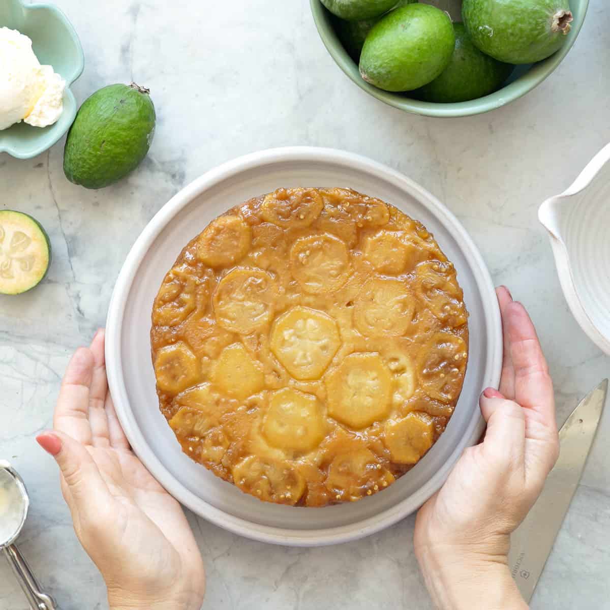 Two hands lifting the plate of the feijoa cake with whole and sliced feijoas and ice cream resting on the bench beside it.