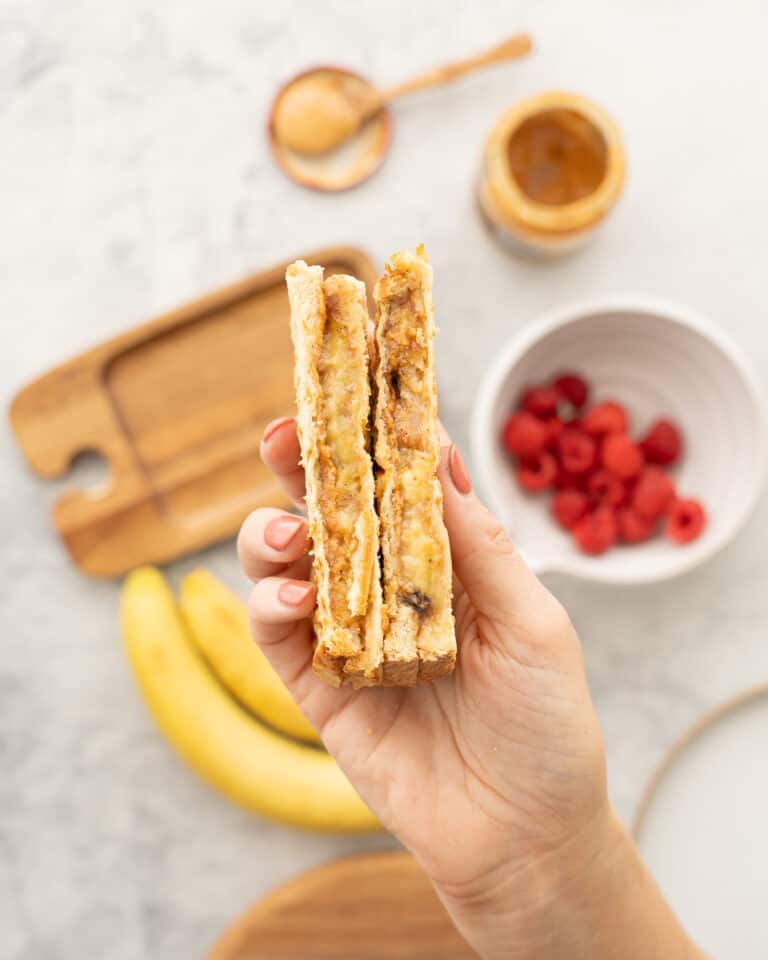 Two grilled peanut butter and banana sandwiches being held up above a chopping board, banana and bowl of raspberries.