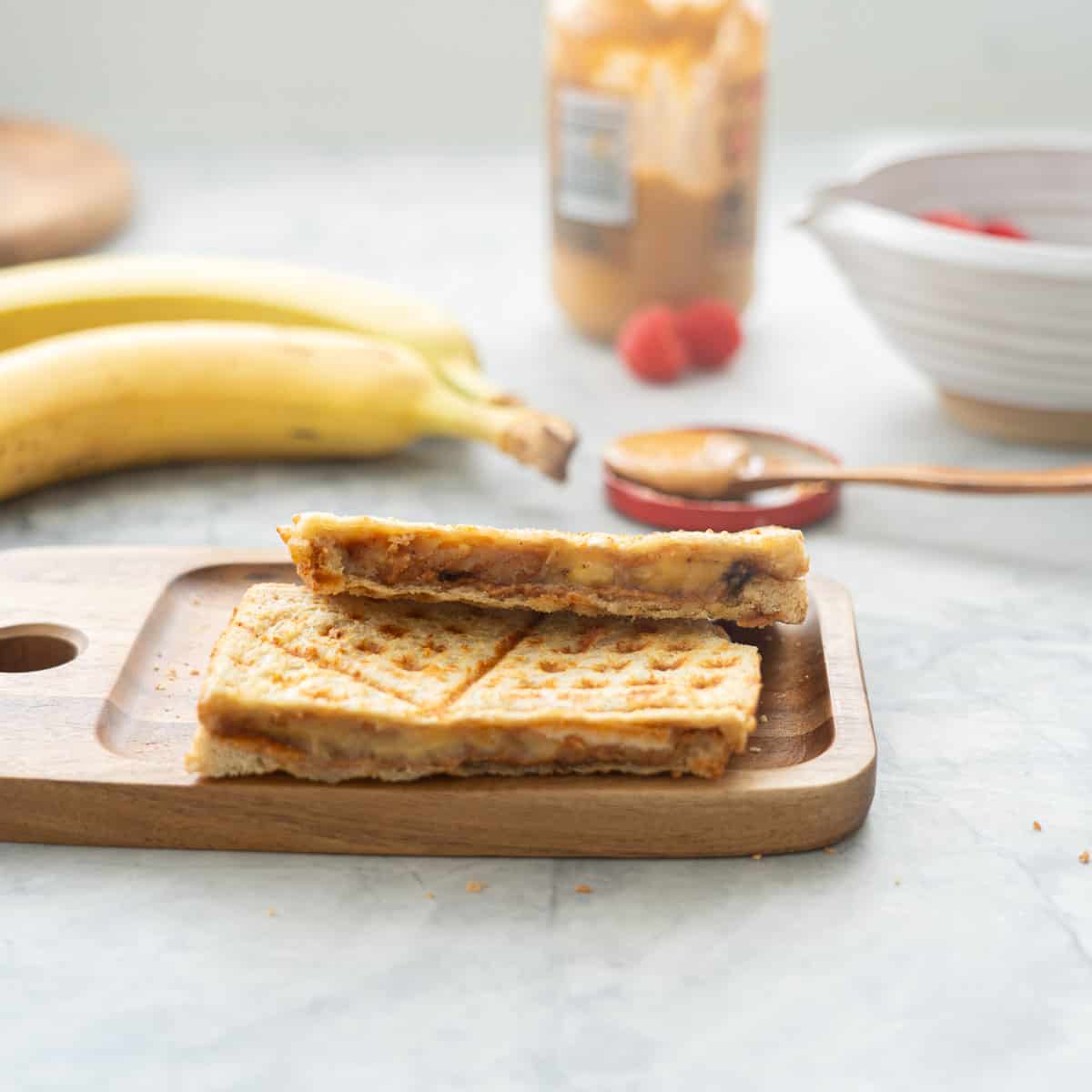 One halved peanut butter banana sandwich resting on a wooden chopping board sitting next to two bananas and a jar of peanut butter