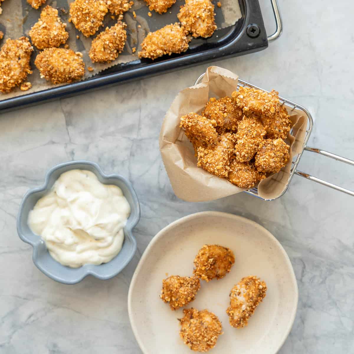 A basket of popcorn chicken, next to a side plate of more chicken pieces and a small ramen filled with aioli.