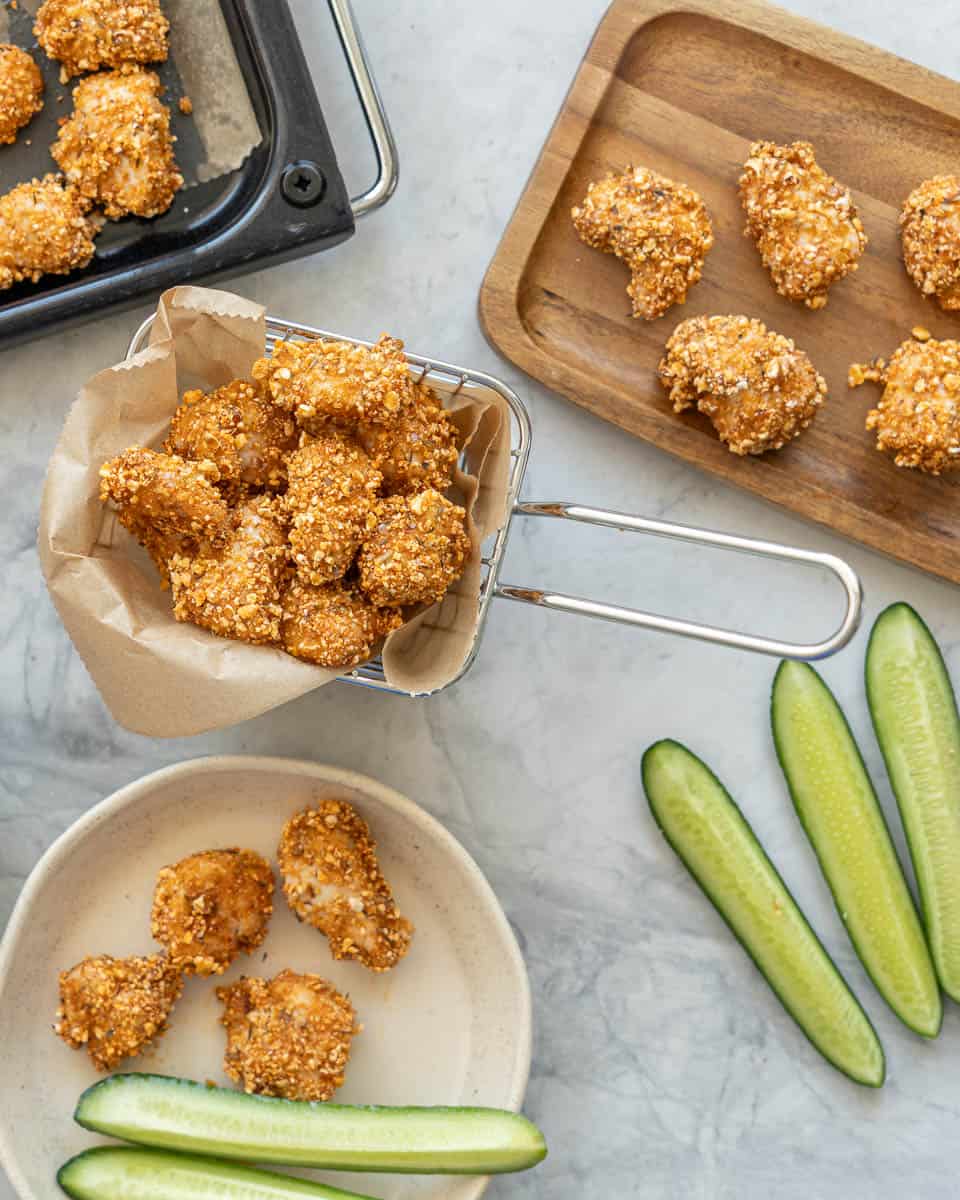 A small mesh basket filled with popcorn chicken, plus a side plate of more popcorn chicken with batons of cucumber.