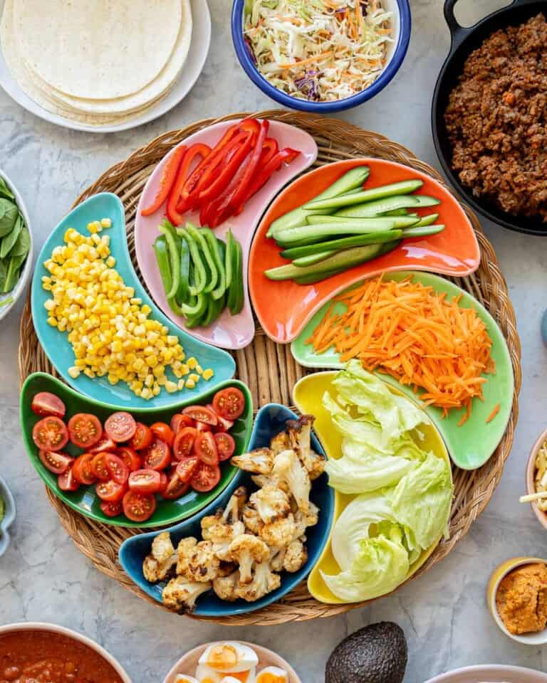 Colourful crescent shaped bowls filled with cut vegetables arranged in a circle on a round wicker tray, with tortillas, mince and beans on a bench top.