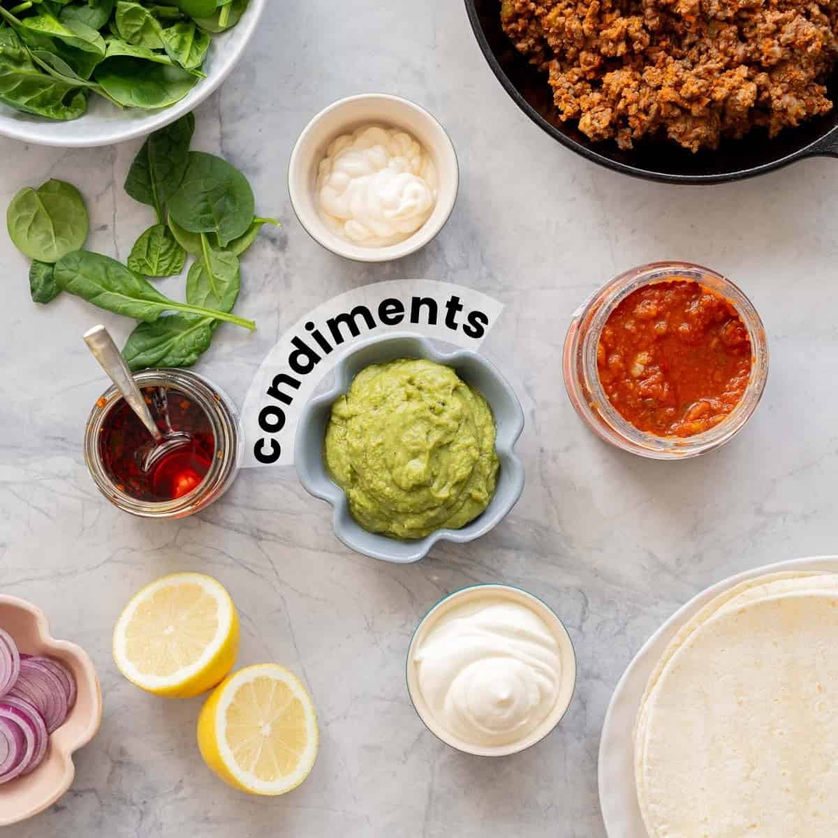 Ramekins and open jars for guacamole, sour cream, salsa, chilli oil and mayonnaise on a bench top with lemon and spinach leaves.