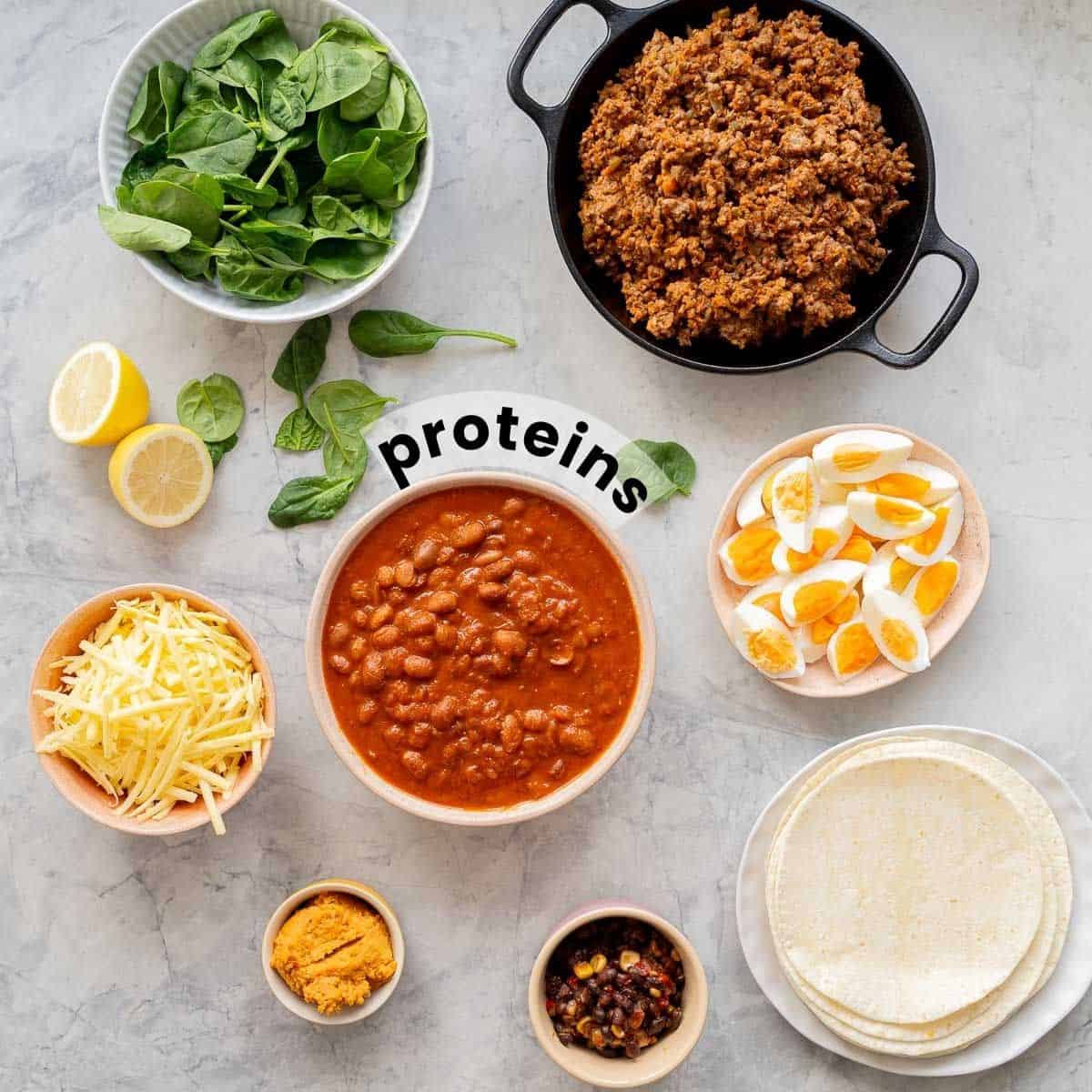 Cooked ground beef, beans, grated cheese, hard boiled eggs and hummus in bowls on a bench top with spinach leaves and lemon halves.