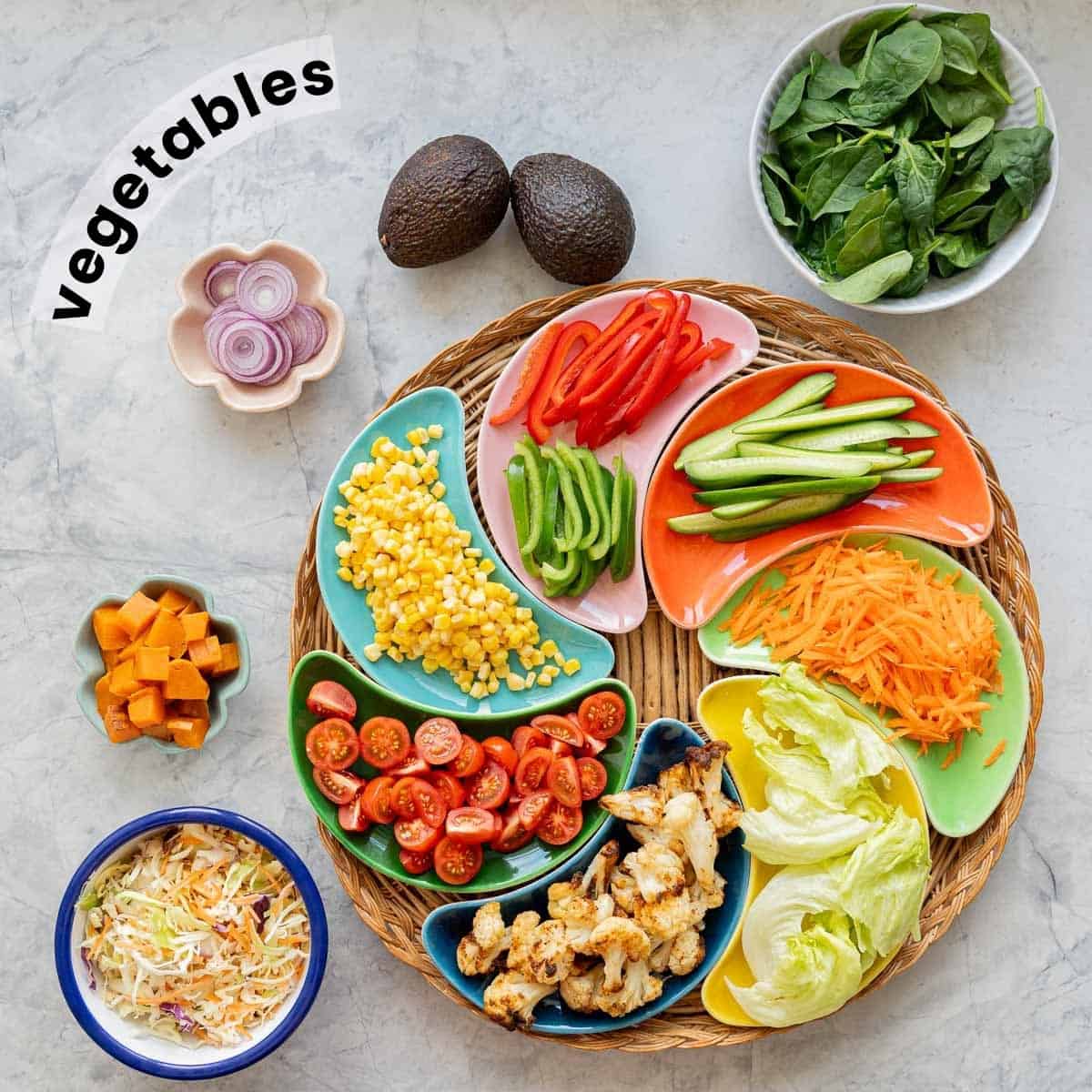 Colourful crescent shaped bowls filled with cut vegetables arranged in a circle on a round wicker tray, with separate bowls of onion slices, spinach leaves, coleslaw and cubed sweet potato on a bench top.