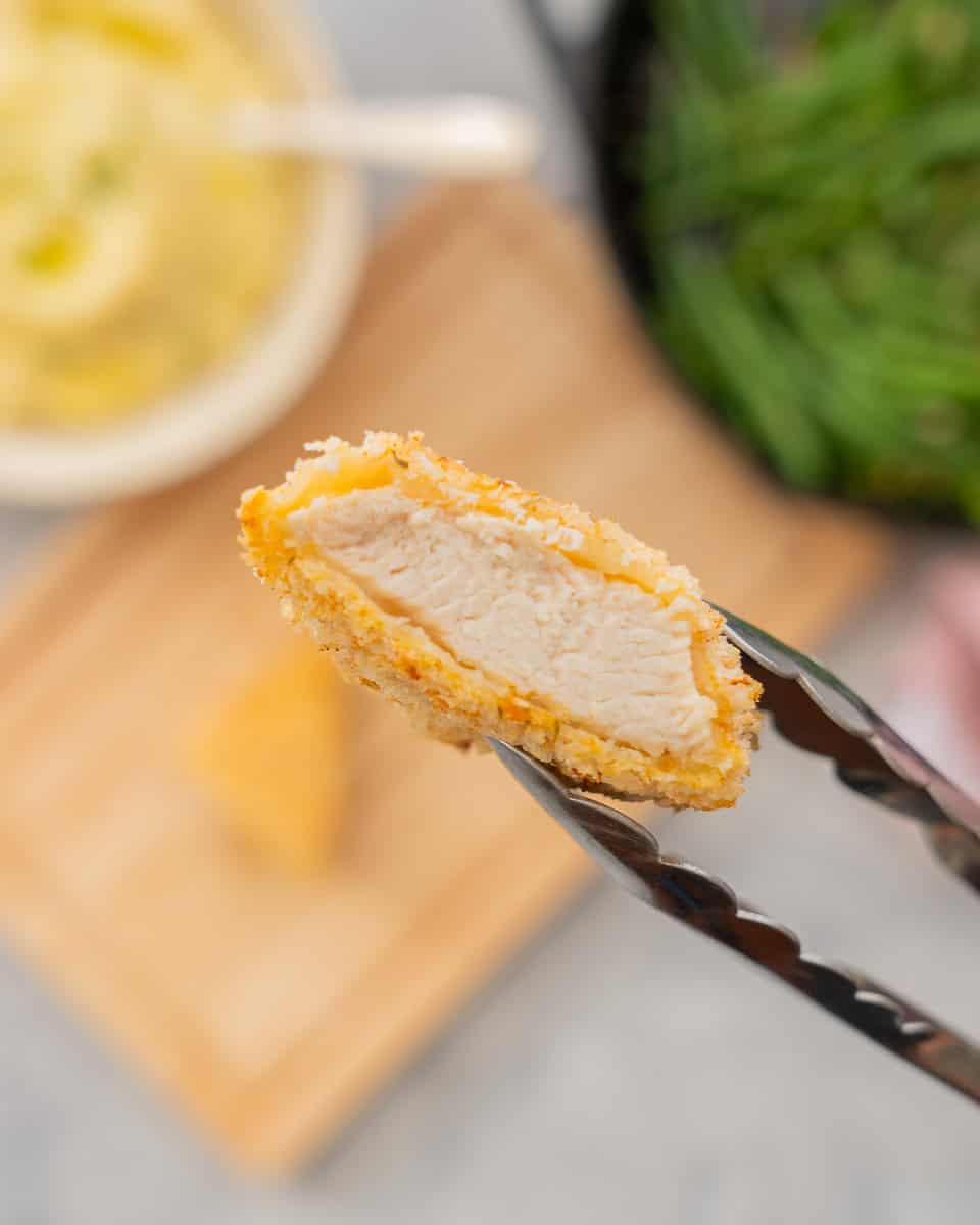 A piece of breaded chicken held in tongs being lifted up to the camera to show the chicken inside the crumb coating.