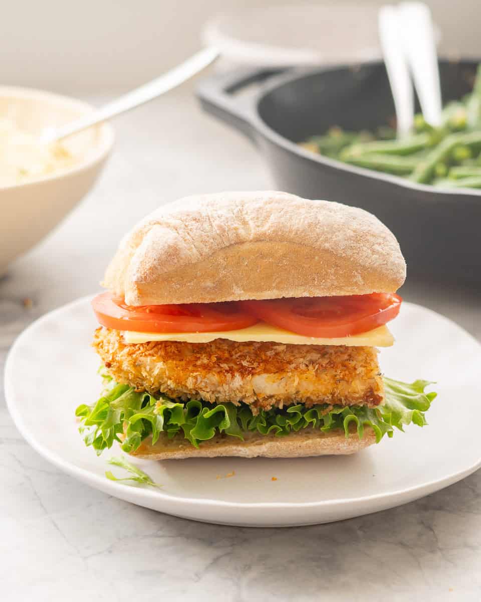 A chicken Birger with tomato slices, cheese, crumbed chicken and lettuce on a small dinner plate.