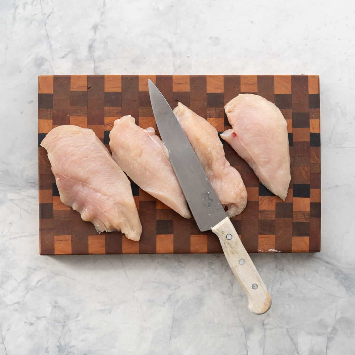 Four chicken cutlets (halved chicken breasts) on a wooden chopping board next to a wooden handled chefs knife.