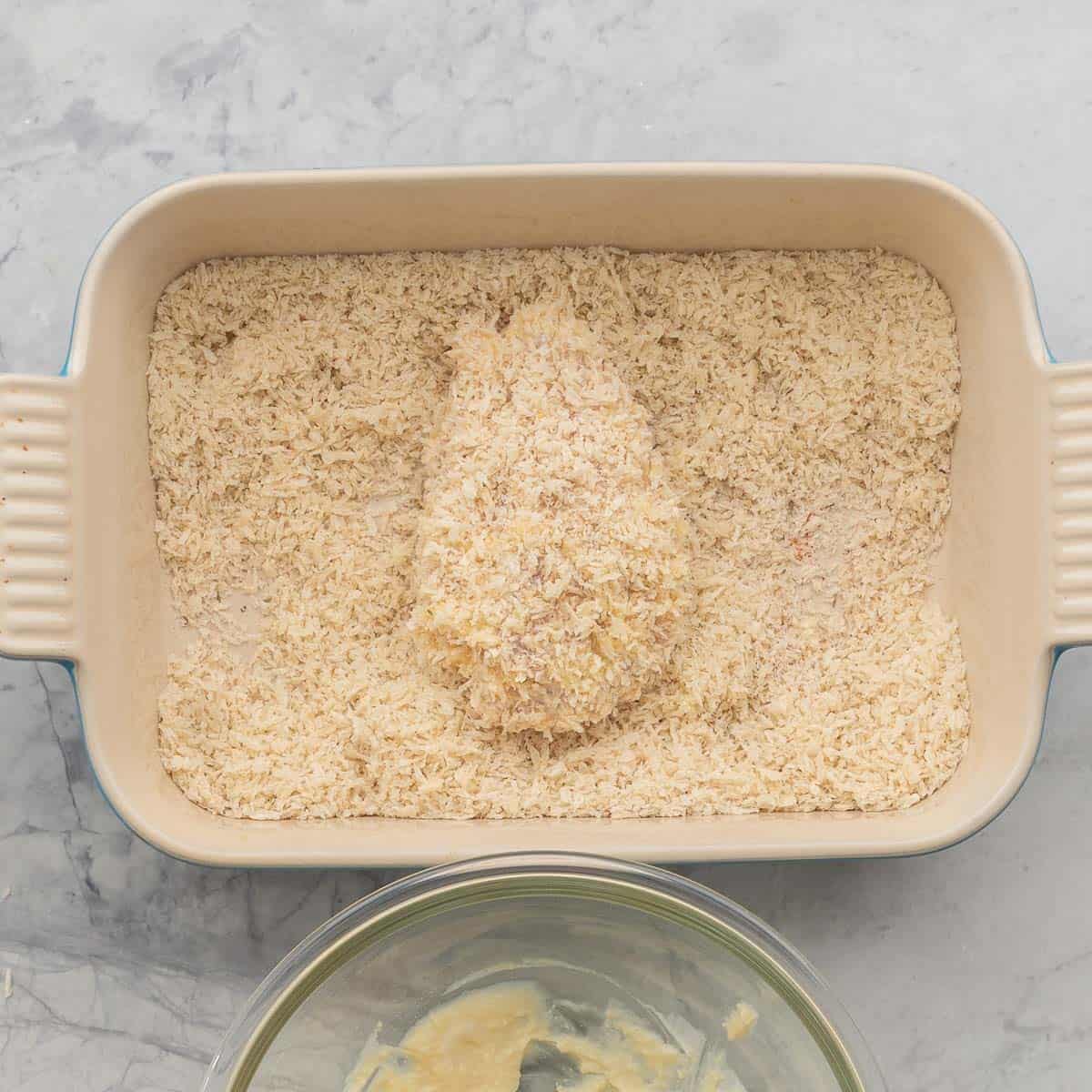 A crumbed chicken cutlet sitting in a rectangular baking dish filled with breadcrumbs.