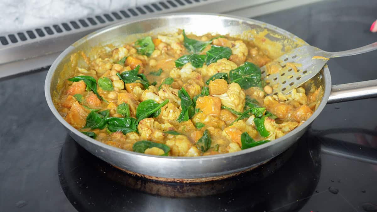 A skillet of curry cooking on a stovetop, chickpeas, cauliflower, spinach and pumpkin pieces visible.