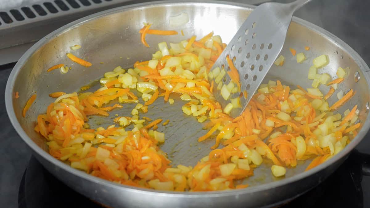 Sautéed onion and grated carrot in a stainless steel fry pan.