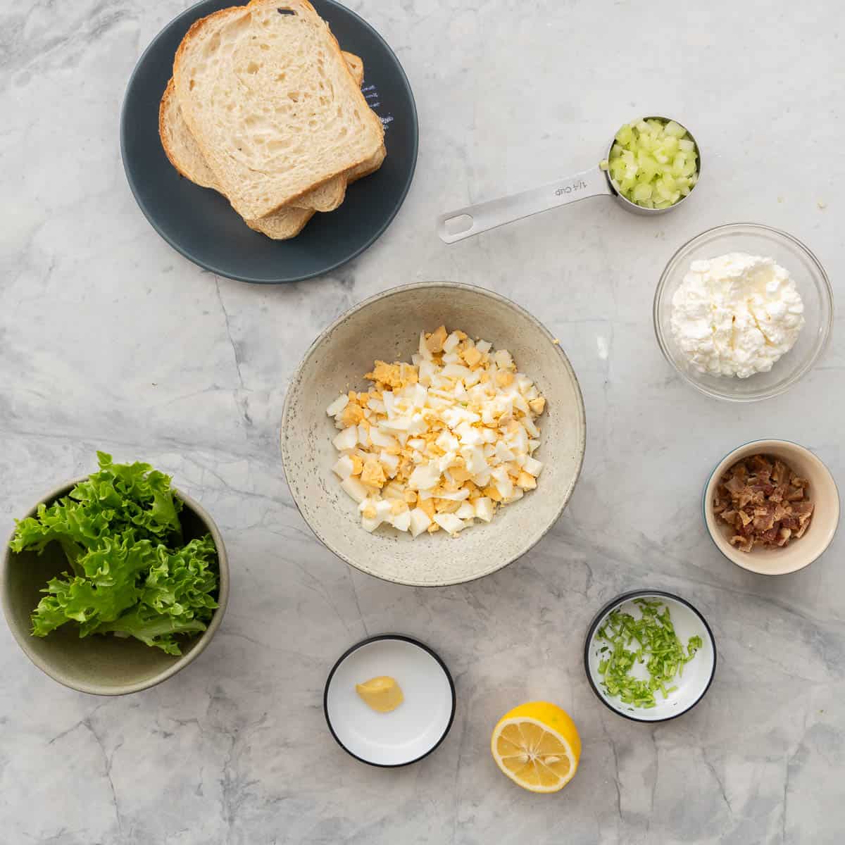 A bowl of chopped hard boiled eggs surrounded by bowls of other egg salad ingredients.