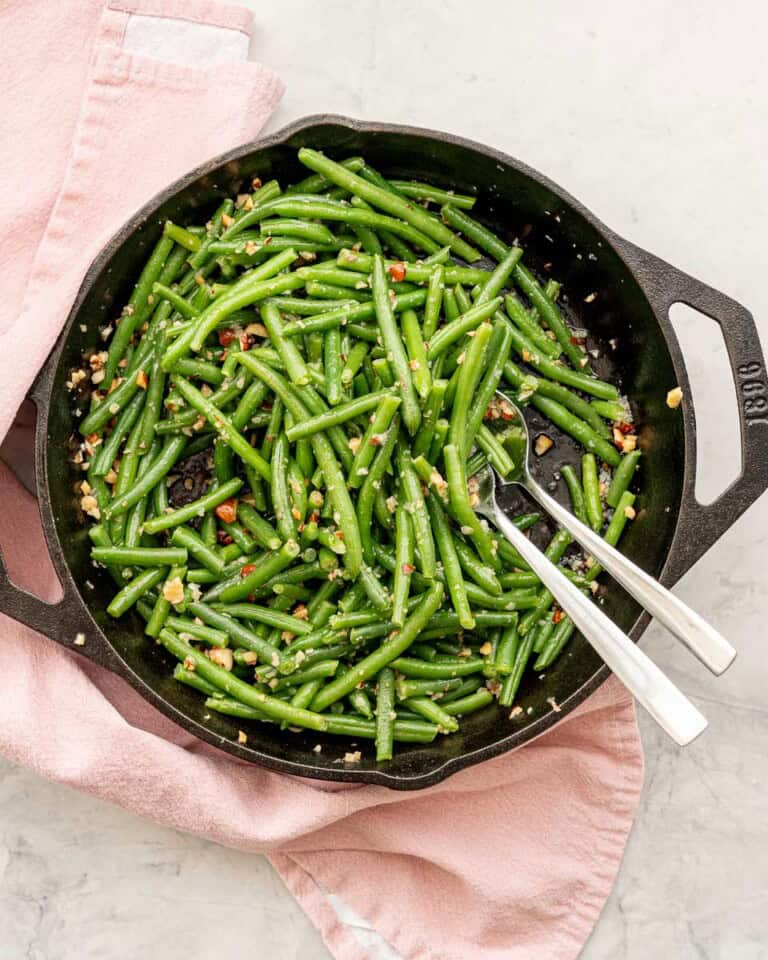 A cast iron skillet of garlic butter coated green beans draped with a pink tea towel sitting on a grey marble bench top.