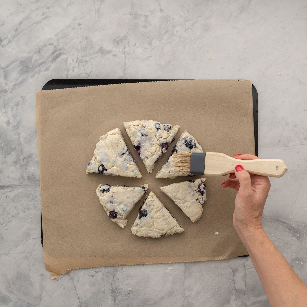 A lined baking tray resting on the bench with wedges of unbaked scone laid out in a circular form with a hand using a brush to apply milk to the top of each.