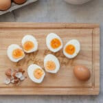 Halved peeled hard boiled eggs on a wooding chopping board next to a pile of egg shell pieces.