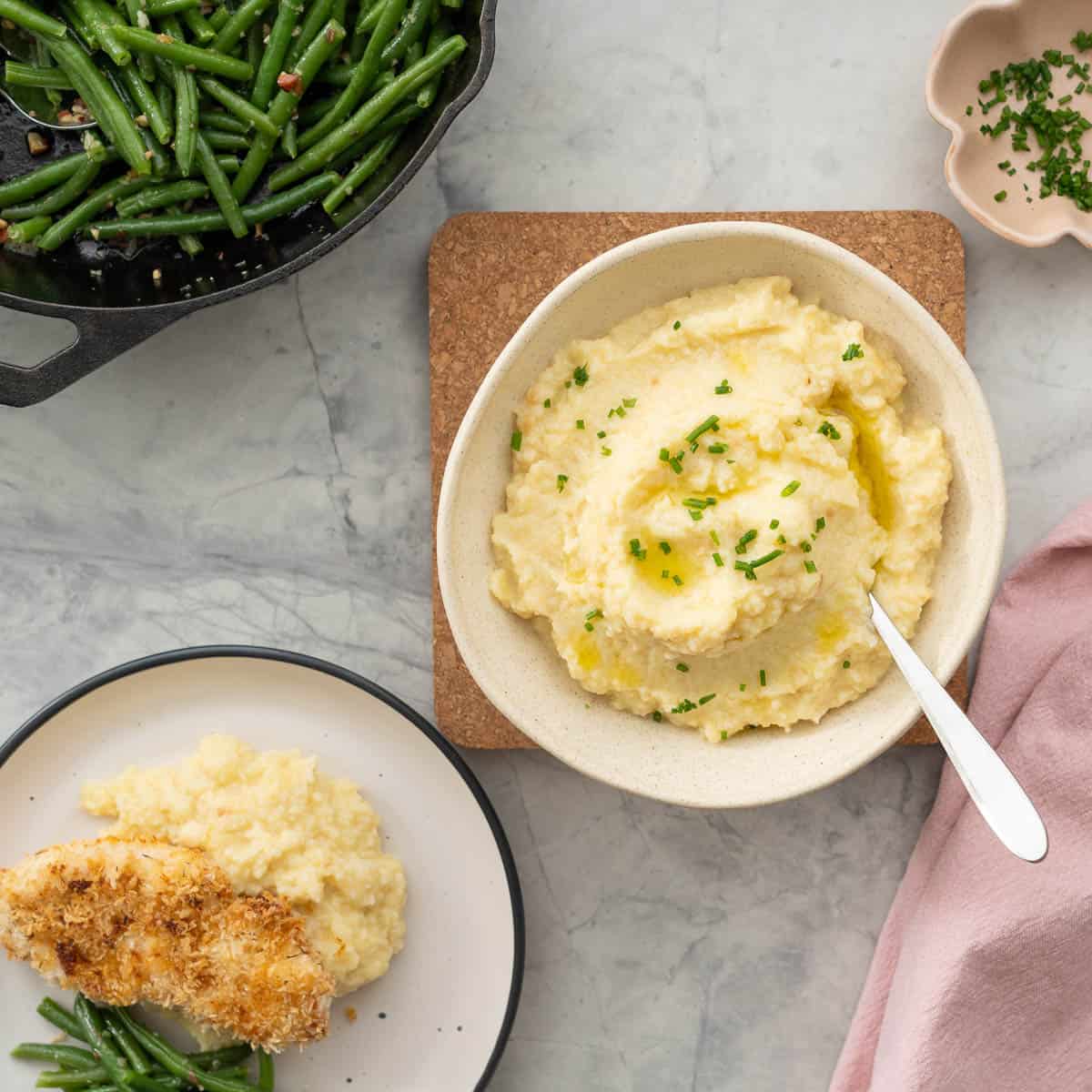 A bowl of potato and cauliflower mash, drizzled with olive oil and sprinkled with chopped chives. next to a dinner plate of crumbed chicken mash and green beans.