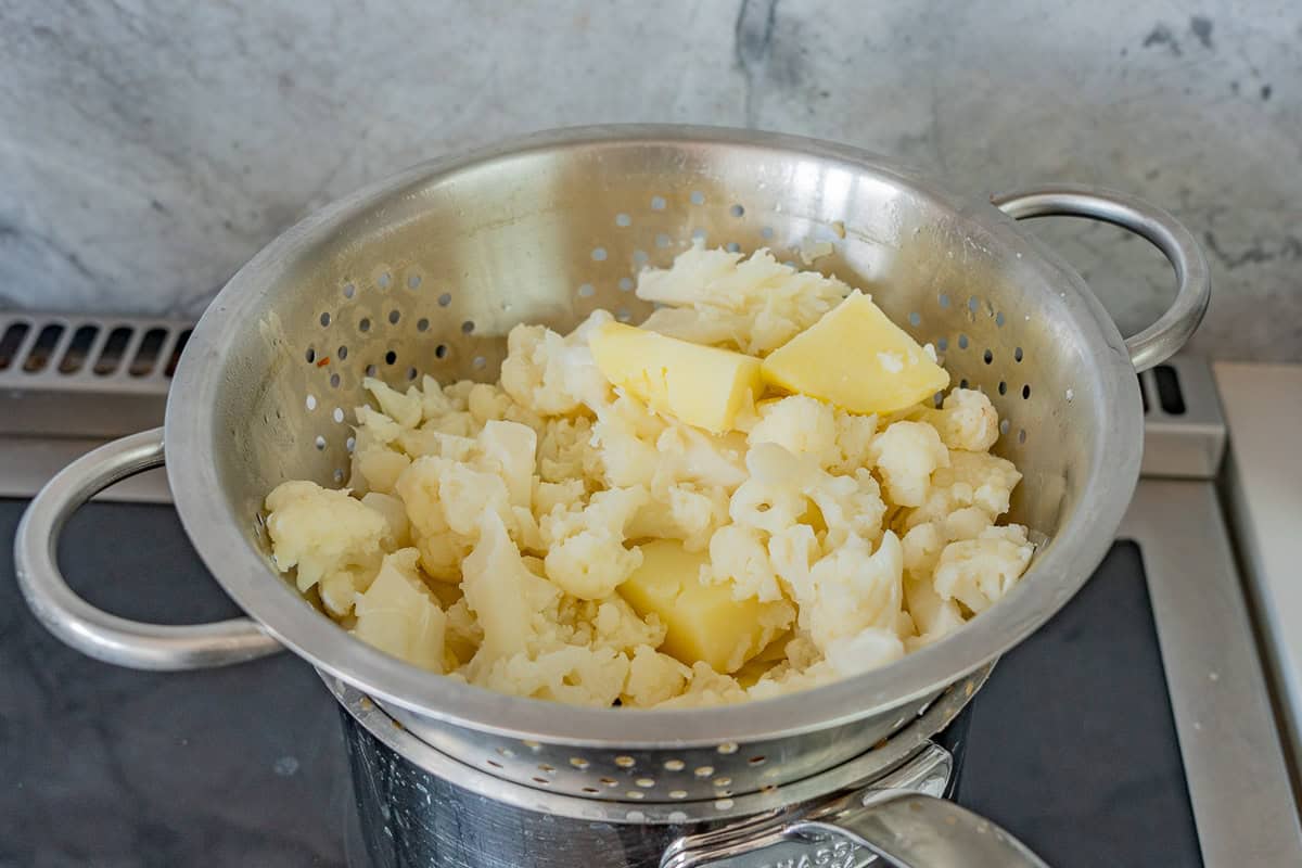 A colander of cooked potato pieces and cauliflower florets draining over a saucepan.