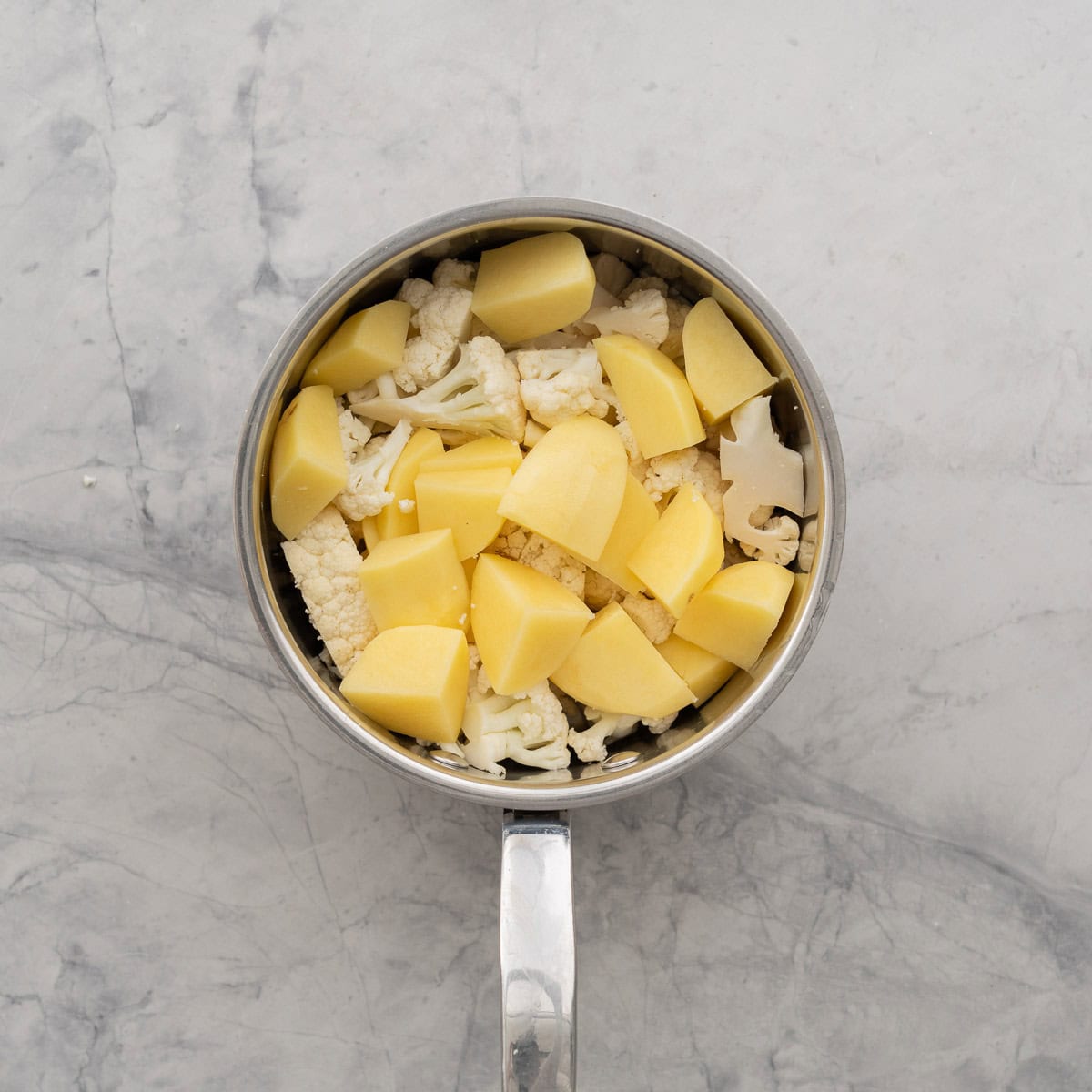 Peeled cubed potato pieces and cauliflower florets in a stainless steel saucepan.