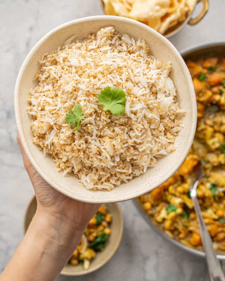 A bowl of cooked rice and quinoa garnished with coriander being held above a skillet of curry.