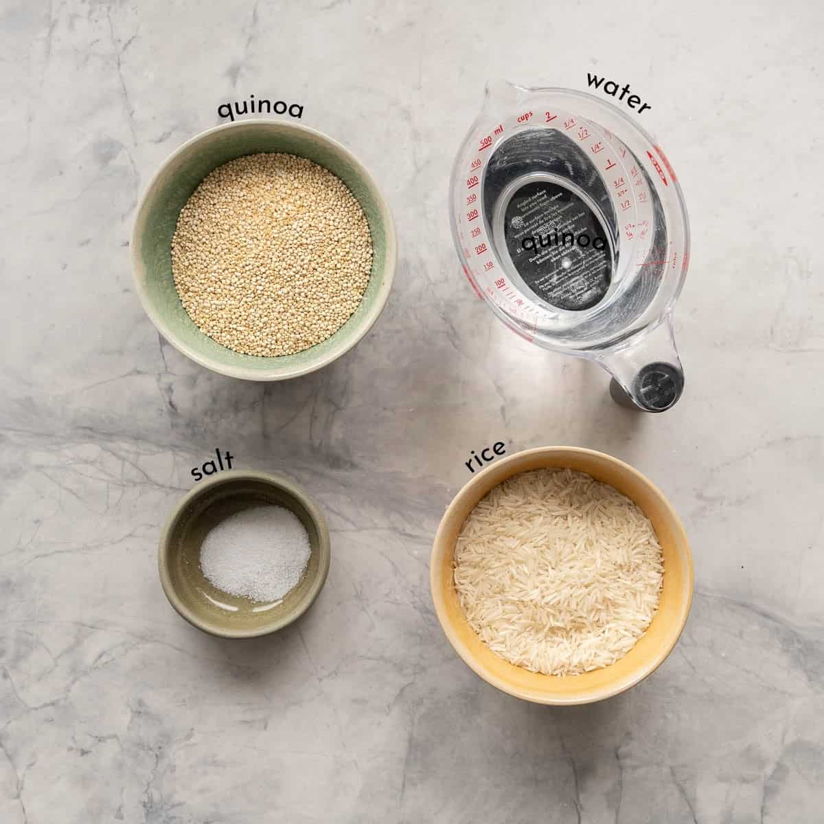 Bowls of quinoa, water, rice and salt each labeled with a text overlay laid out on a bench top.