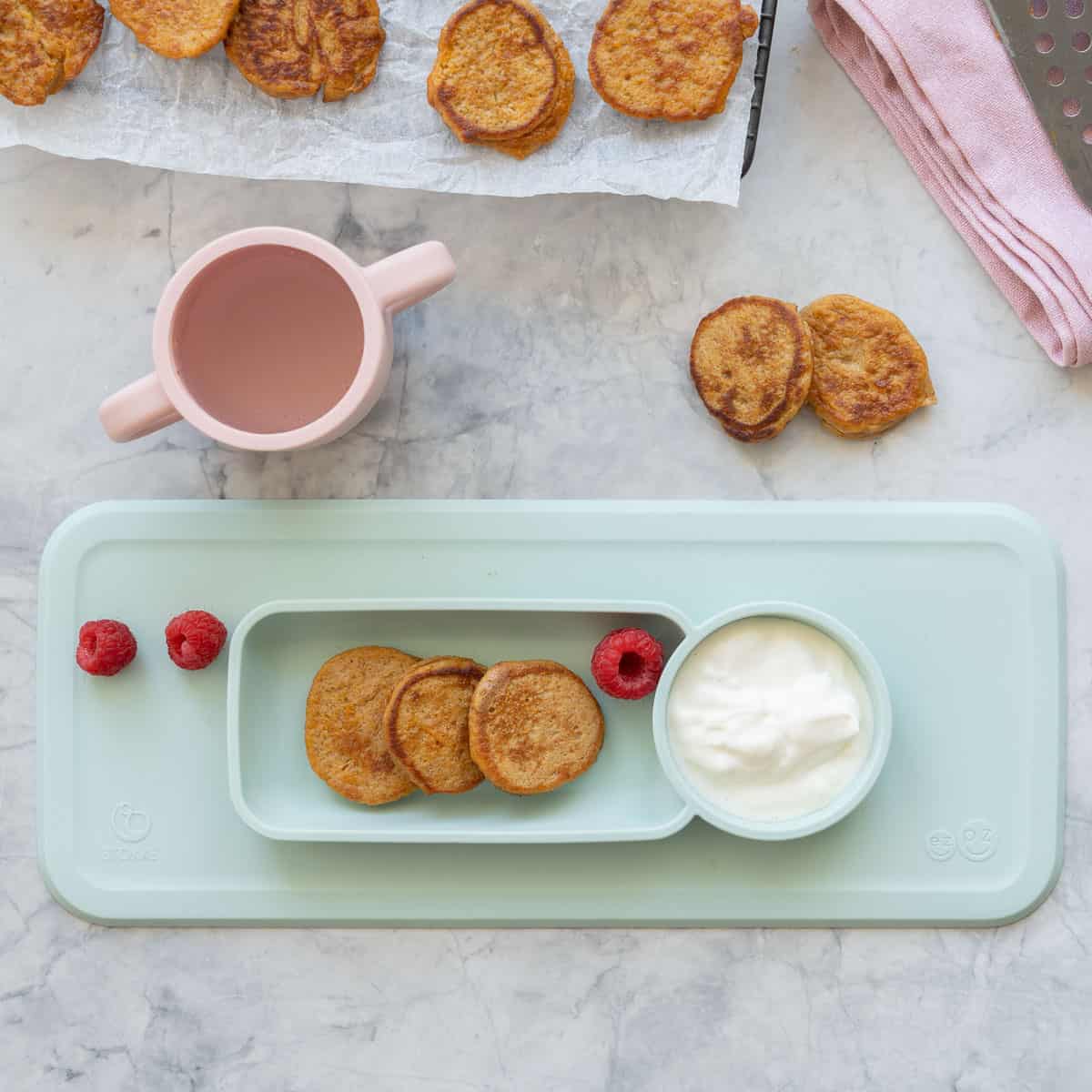 A baby led weaning plate of pancakes yogurt and raspberries next to a small pink sippy cup.
