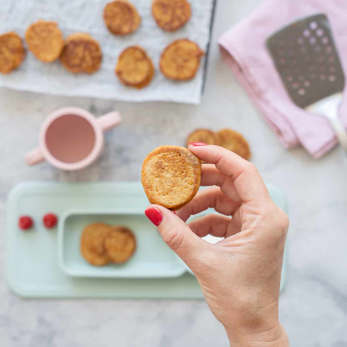 A small pancake being held up to the camera above a baby led weaning plate of more pancakes, yogurt and raspberries.