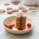 A stack of seven pancakes drizzled with maple, the stream of maple syrup is visible on a side plate with two raspberries.