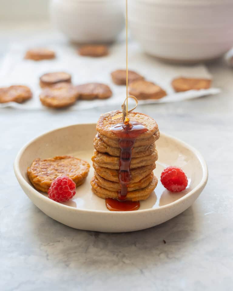 A stack of seven pancakes drizzled with maple, the stream of maple syrup is visible on a side plate with two raspberries.