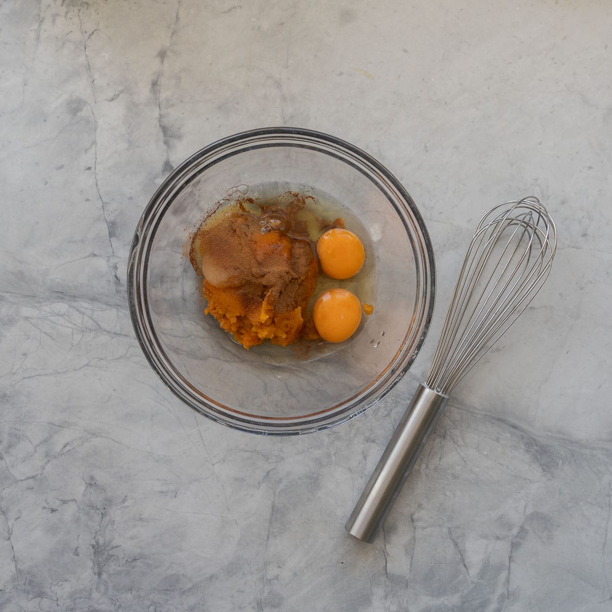 Eggs, mashed sweet potato and ground cinnamon in a glass mixing bowl with a whisk sitting next to the bowl.