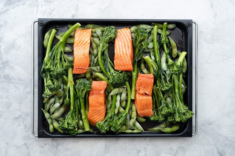 Four pieces of fresh salmon on top of vegetables on baking tray on benchtop.