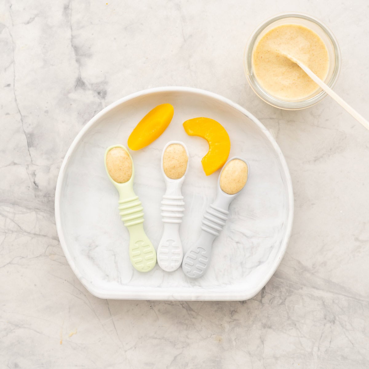 A baby plate holding 3 baby spoons pre-filled with peach chia pudding and pieces of tinned peach.