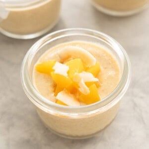 A peach coloured pudding in a glass jar topped with glistening cubes of peach and toasted coconut chips.