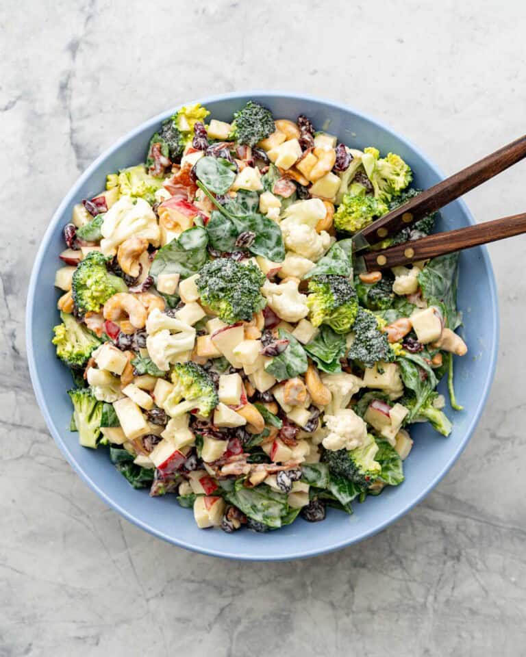 A big blue salad bowl filled with apple spinach broccoli cauliflower raisins and cashews, topped with creamy salad dressing, with wood tongs in bowl.
