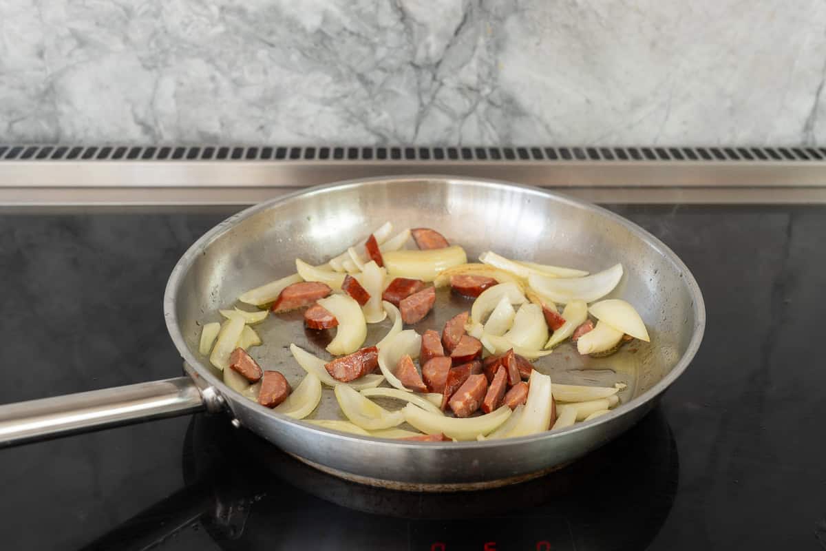 A large fry pan on the cooktop sauteing onions and chorizo in oil.