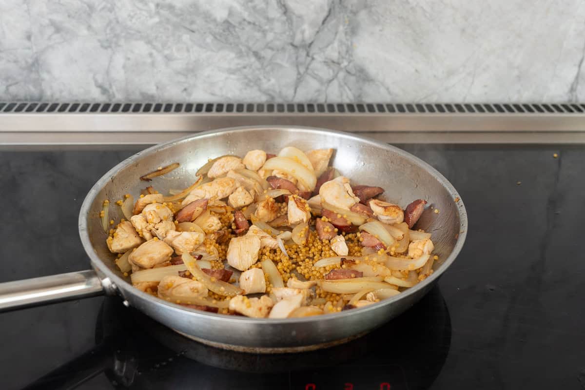 A large fry pan on the cooktop with onions, chorizo, chicken and couscous simmering.