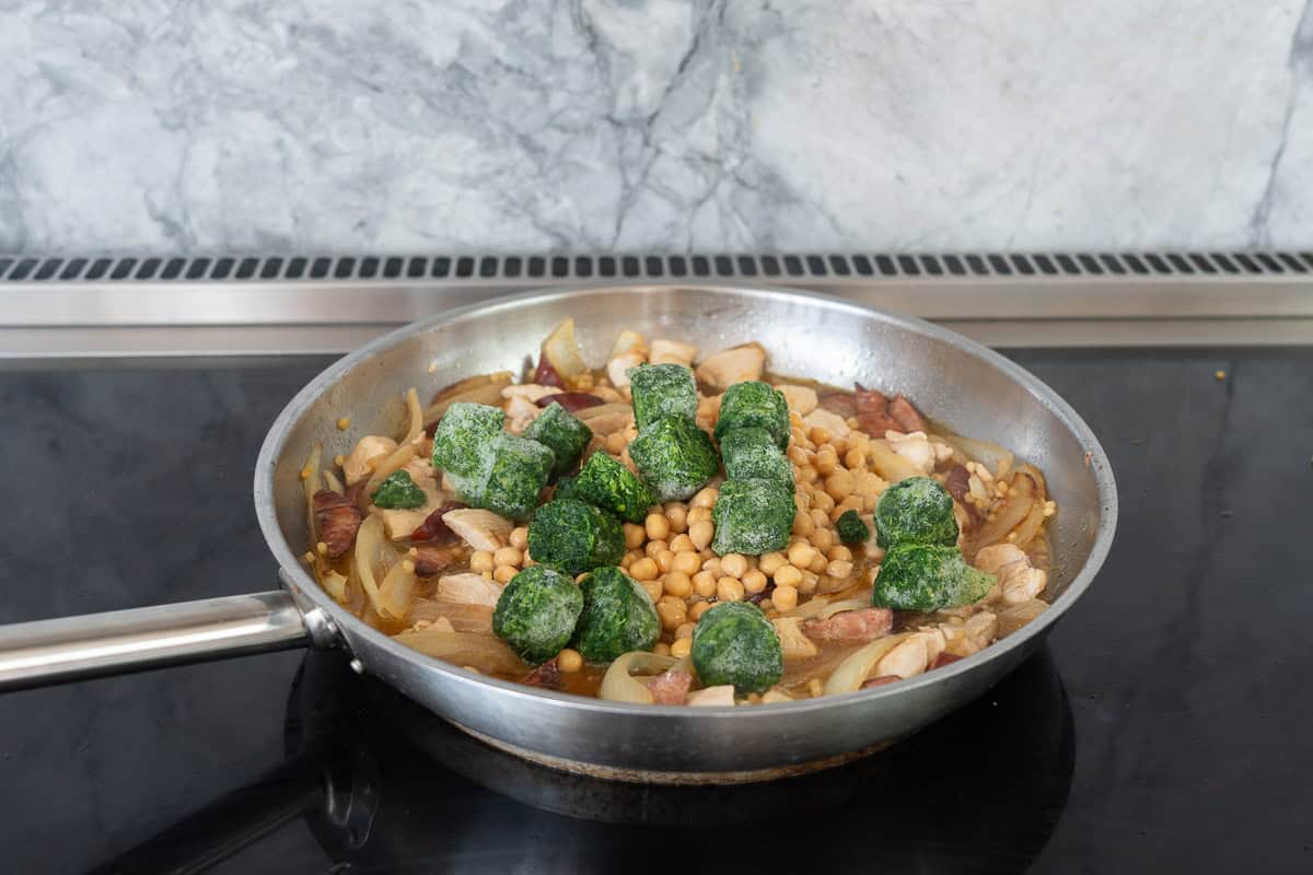 A large fry pan on the simmering onions, chorizo, chicken and couscous with frozen cubes of spinach added.