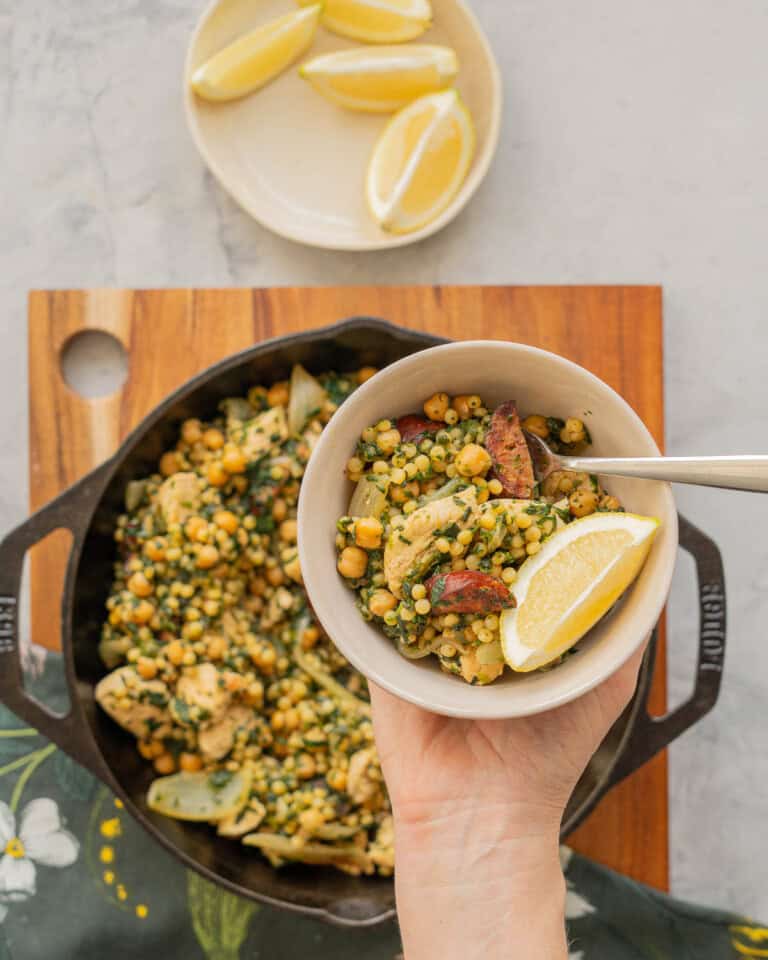 A large cast iron skillet of Chicken Couscous resting on a wooden chopping board on a bench with a hand holding a serving on a plate above it.