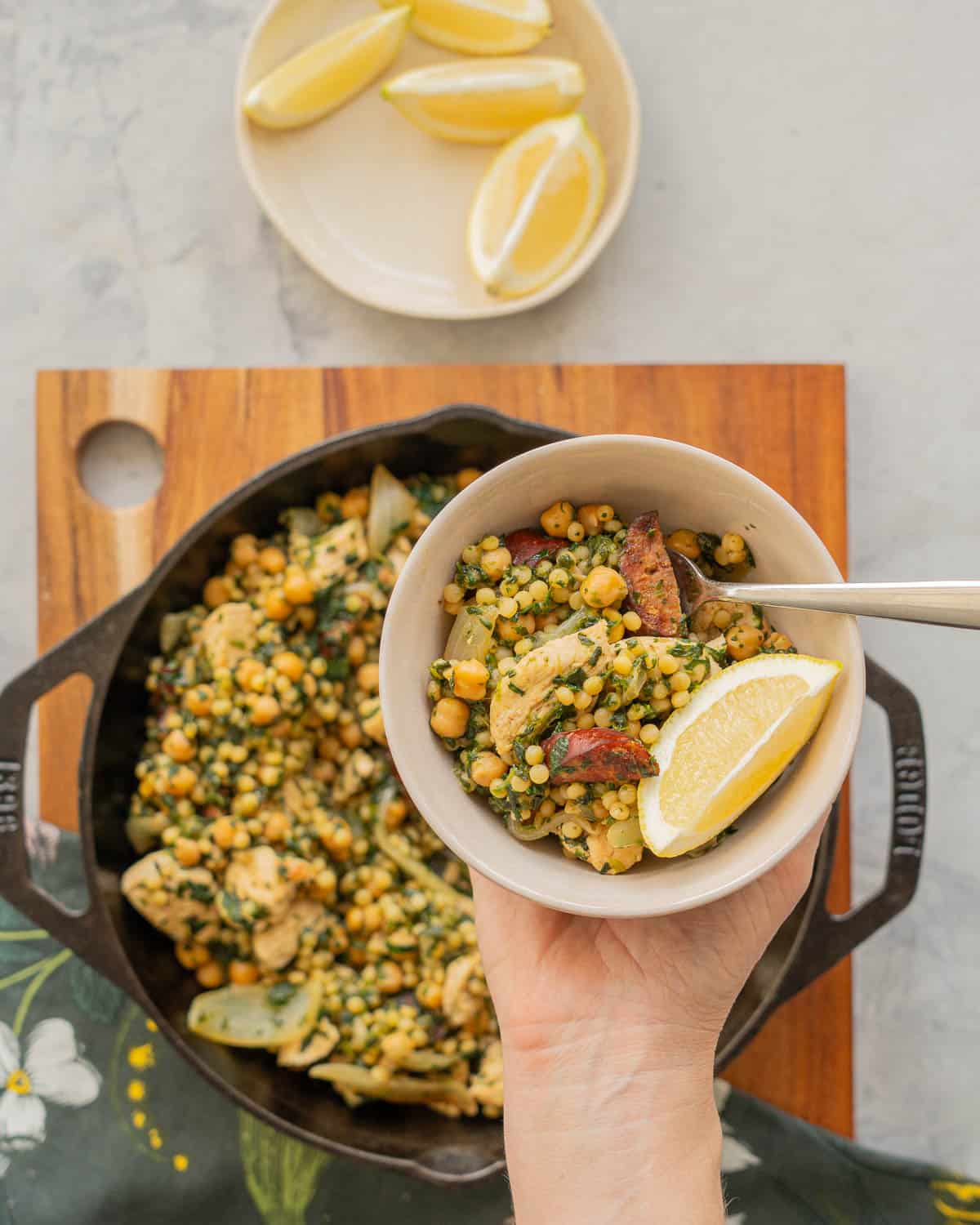 A large cast iron skillet of Chicken Couscous resting on a wooden chopping board on a bench with a hand holding a serving on a plate above it.