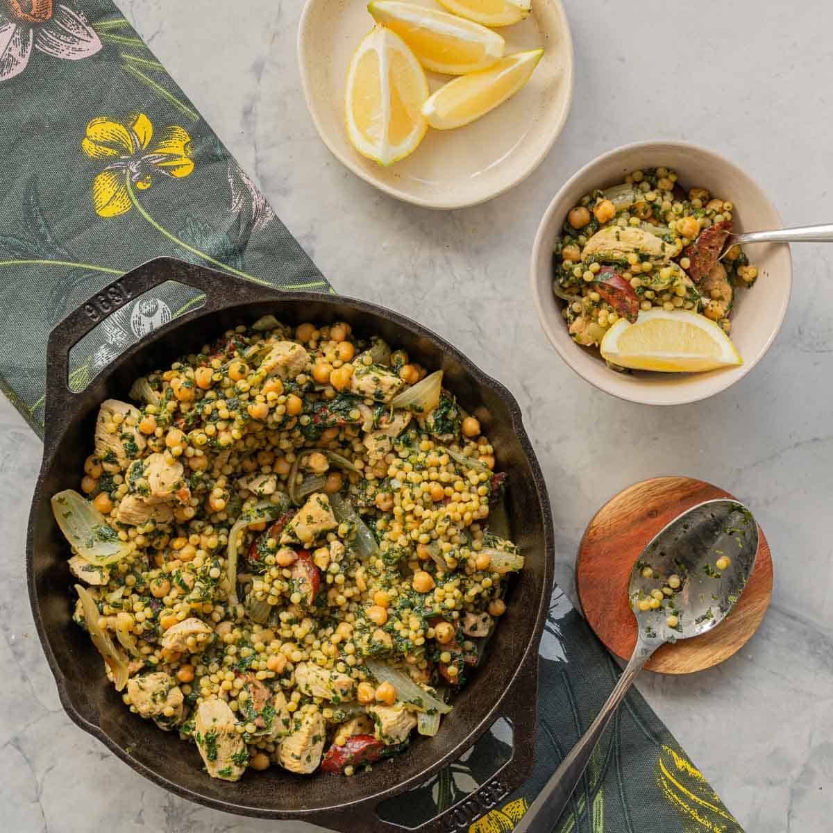 A skillet of chicken couscous and chorizo on a green placemat next t a side plate of lemon wedges.