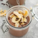 A chocolate pudding in a jar topped with banana slices, coconut chips and chocolate chips.