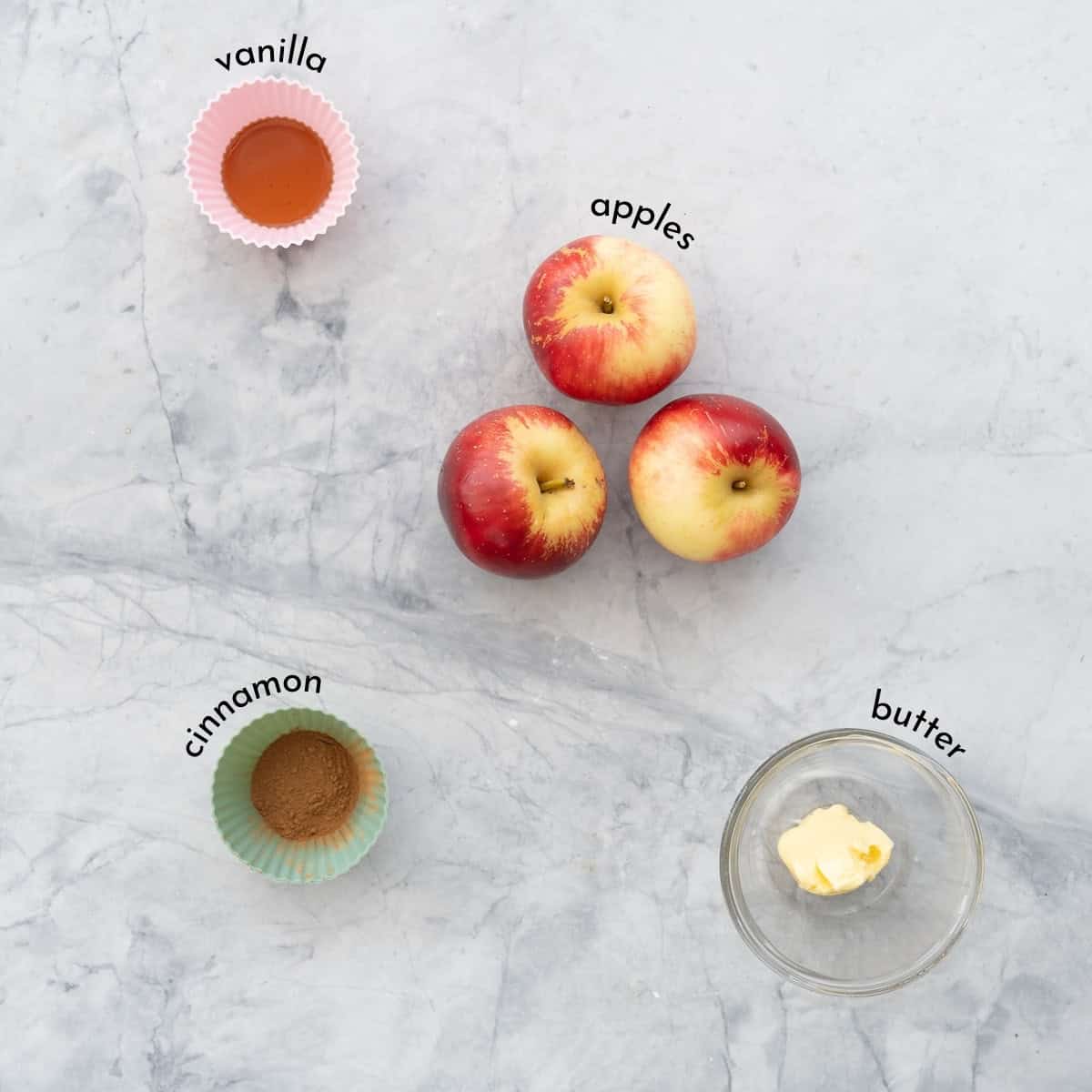 Ramekins of vanilla extract, cinnamon, butter and three loose apples on a bench top with text overlay ingredient labels.