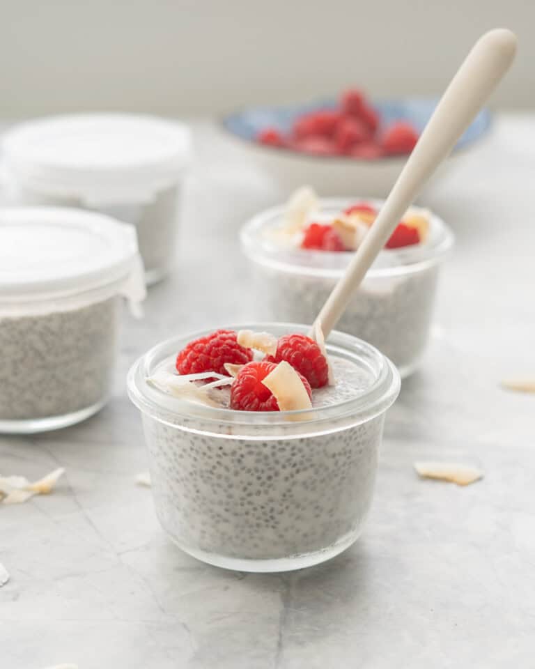 A small glass container of coconut chia pudding topped with raspberries and coconut flakes, wood spoon in bowl.