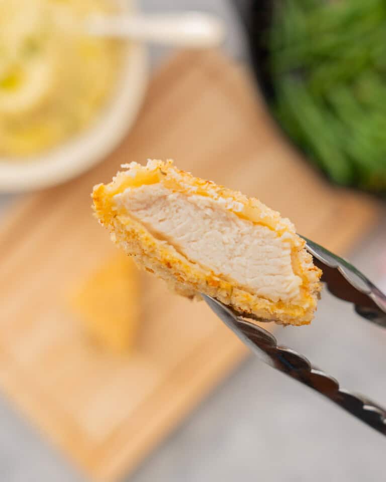 A piece of breaded chicken held in tongs being lifted up to the camera to show the chicken inside the crumb coating.