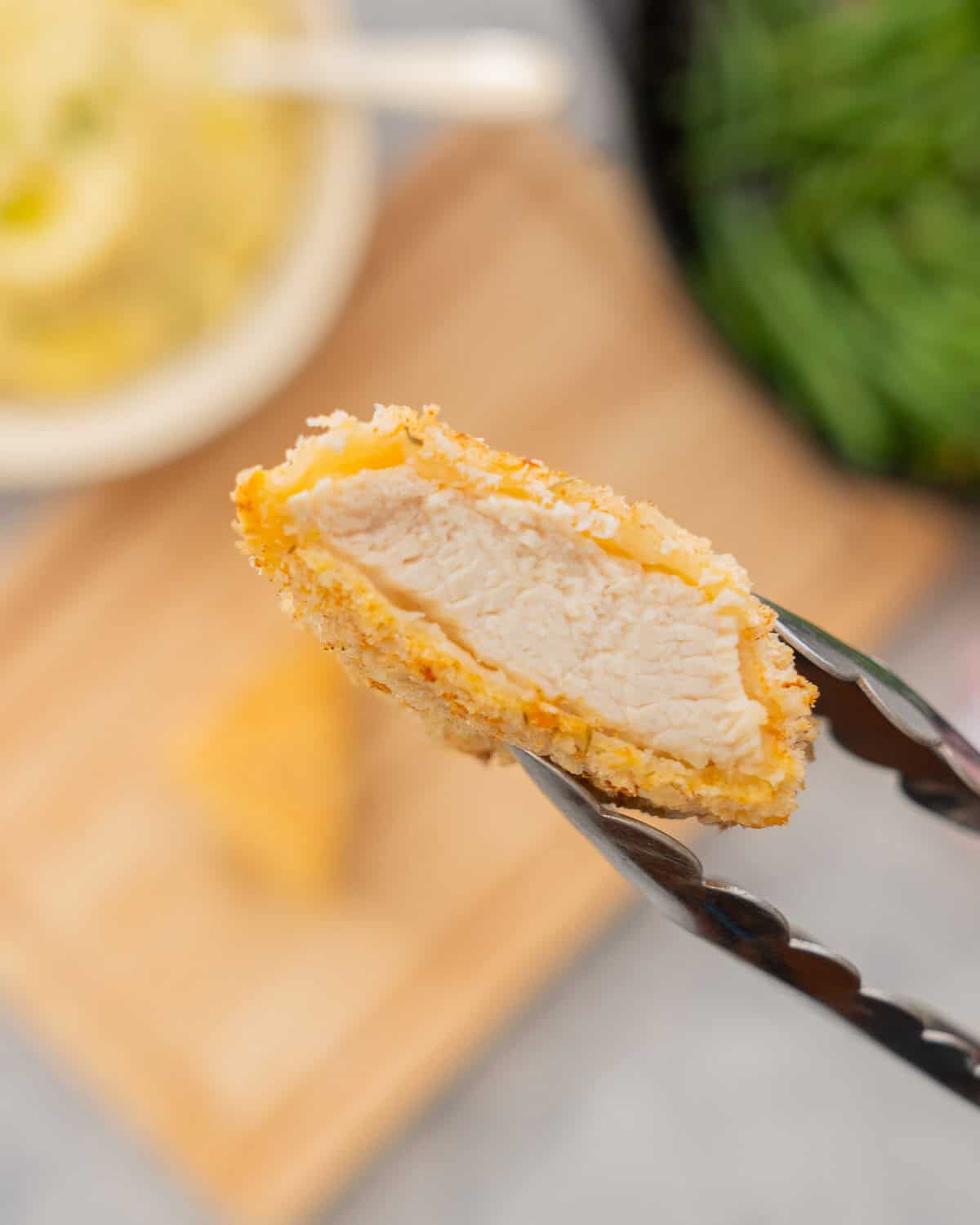 A piece of breaded chicken held in tongs being lifted up to the camera to show the chicken inside the crumb coating.