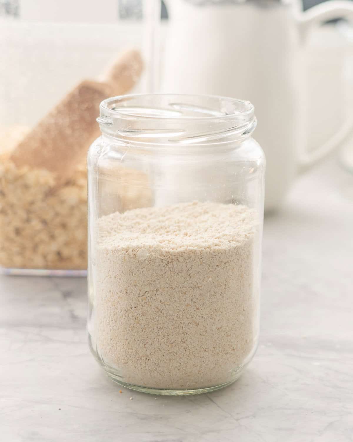 One jar of oat flour resting on the bench in front of a container of oats.