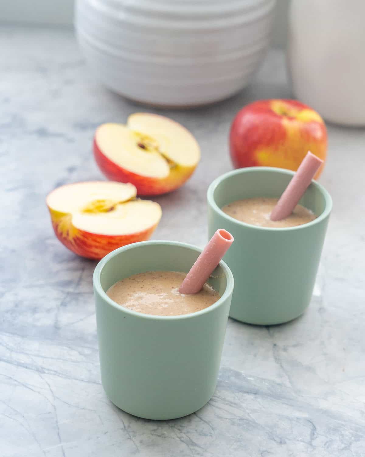 Two small green toddler cups with pink silicone straws filled with pale brown smoothie, apple pieces in the background.