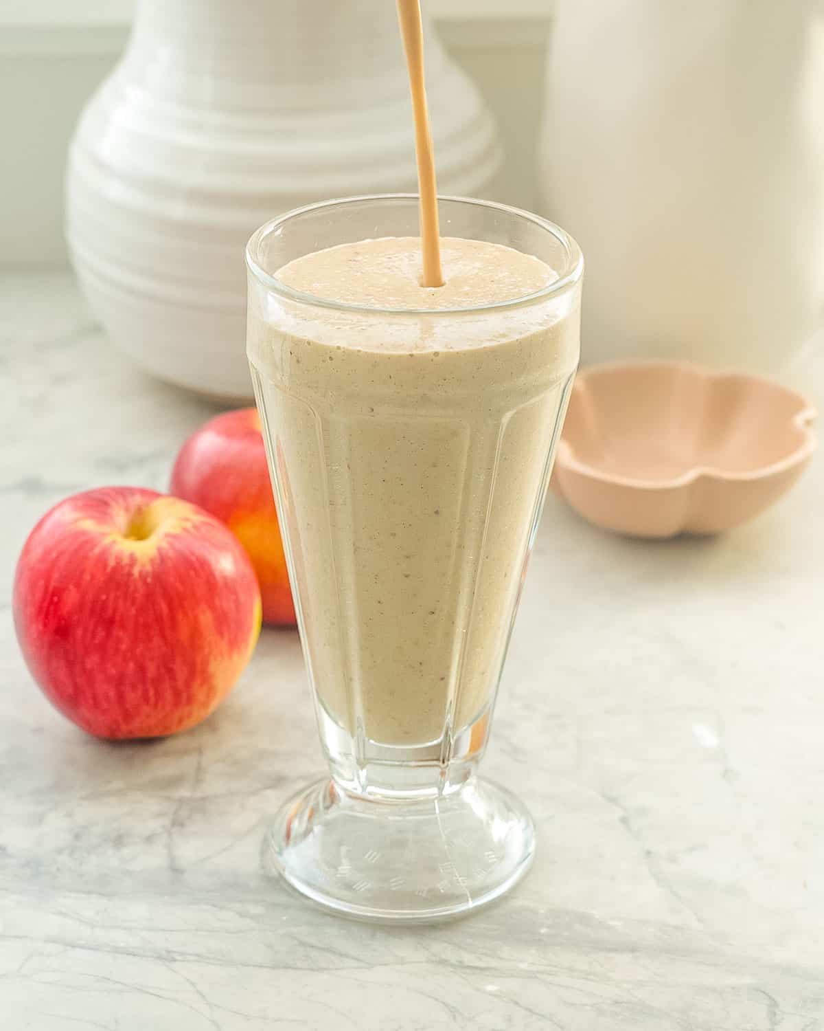 A tall sundae glass almost fill with smoothie, a thin stream of smoothie pouring in still visible, two apples and white ceramic jug behind the sundae glass.